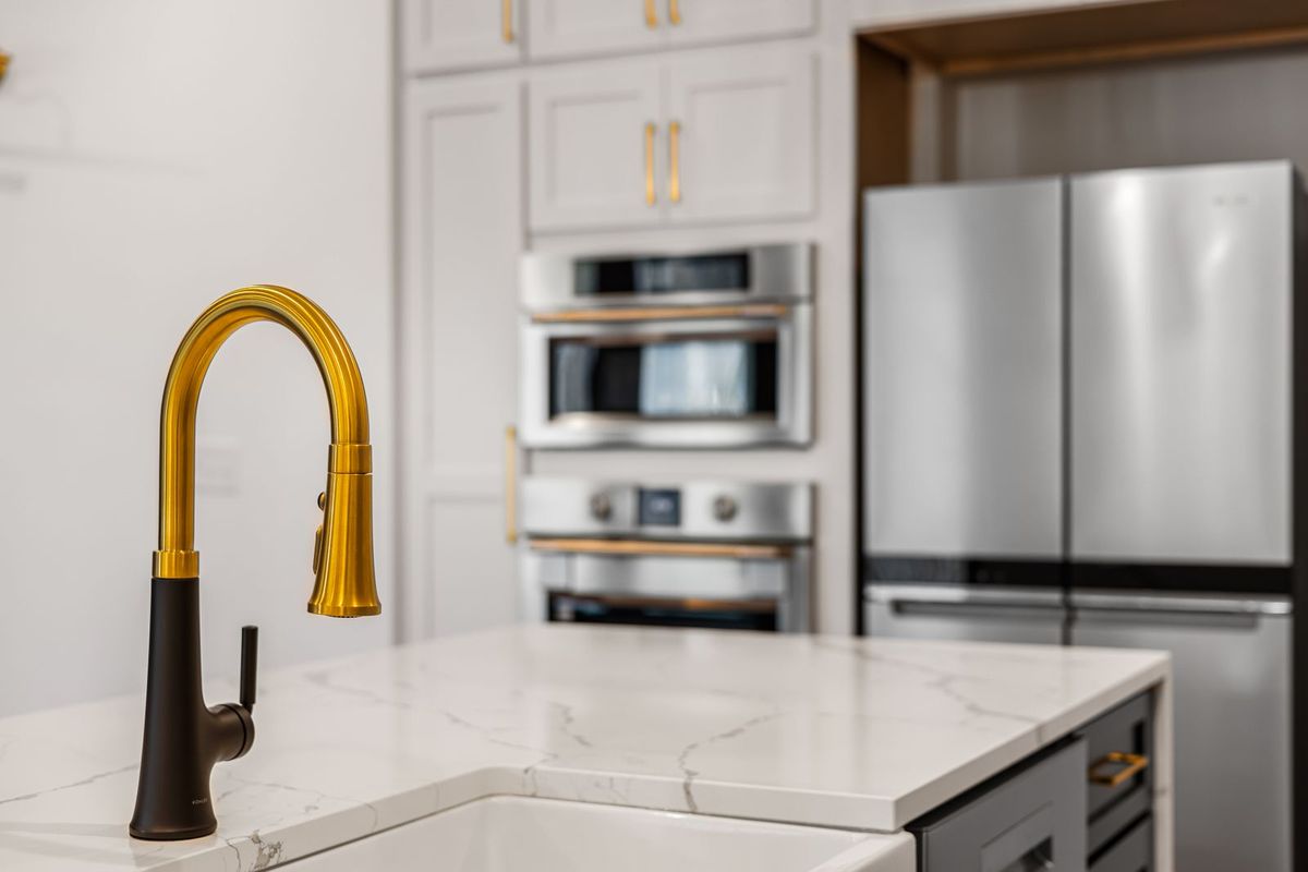 Brass kitchen faucet on a white quartz island with built-in wall ovens behind
